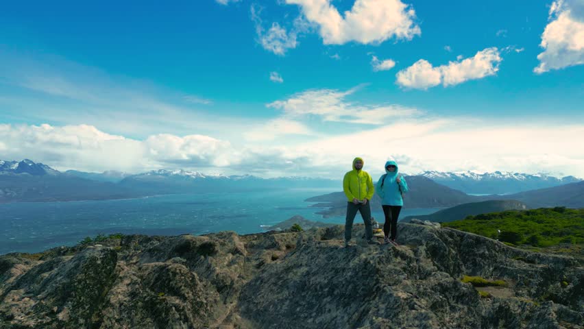 A couple woman and man stand on mountain peak overlooking the Beagle Channel at windy day in Ushuaia, Tierra del Fuego, Argentina. Aerial drone video.
