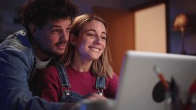 Two happy young Caucasian people together looking at computer sitting at desk in living room at night. Smiling millennial affectionate couple at home using laptop. Domestic relations and leisure time - Powered by Shutterstock - Get 15% off with code: PIKWIZARD15