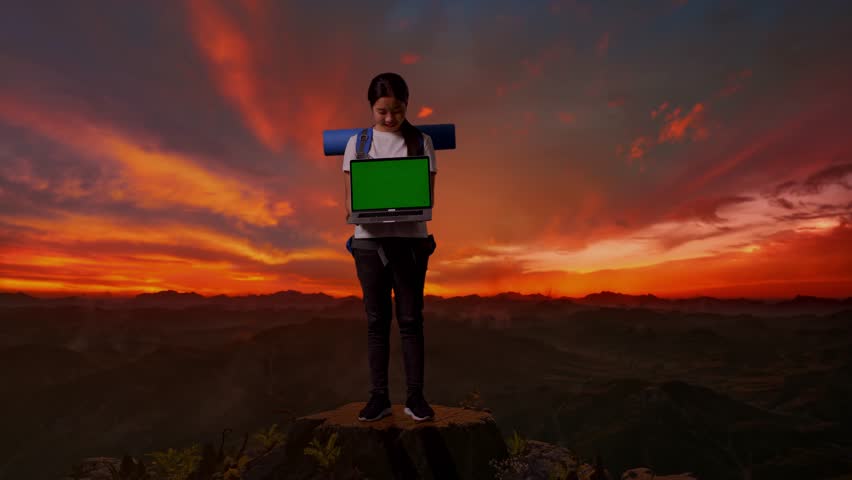 Full Body Of Asian Female Hiker With Mountaineering Backpack Smiling And Showing Mock Up Green Screen Laptop While Standing On The Top Of Mountain During Sunset Time