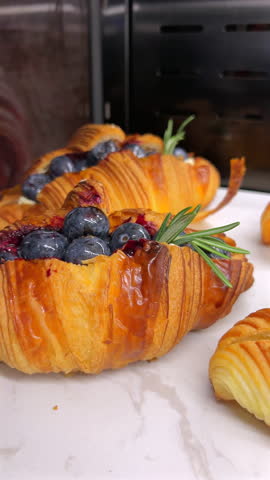 Freshly baked croissants adorned with blueberries and sweet glaze, garnished with rosemary, on kitchen countertop with oven in background. Gourmet pastry and breakfast.