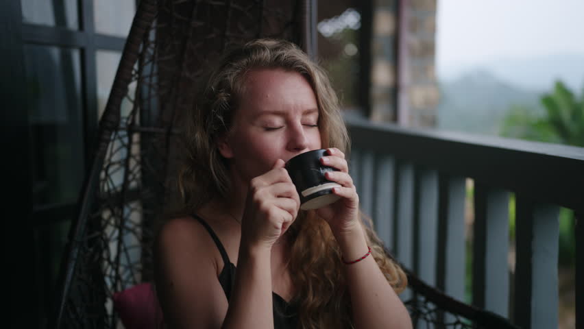Woman sips tea in rope swing chair on luxurious balcony with breathtaking mountain scenery, surrounded by tea gardens. Relaxed travel moment, leisure retreat with serene nature view. Slow motion.