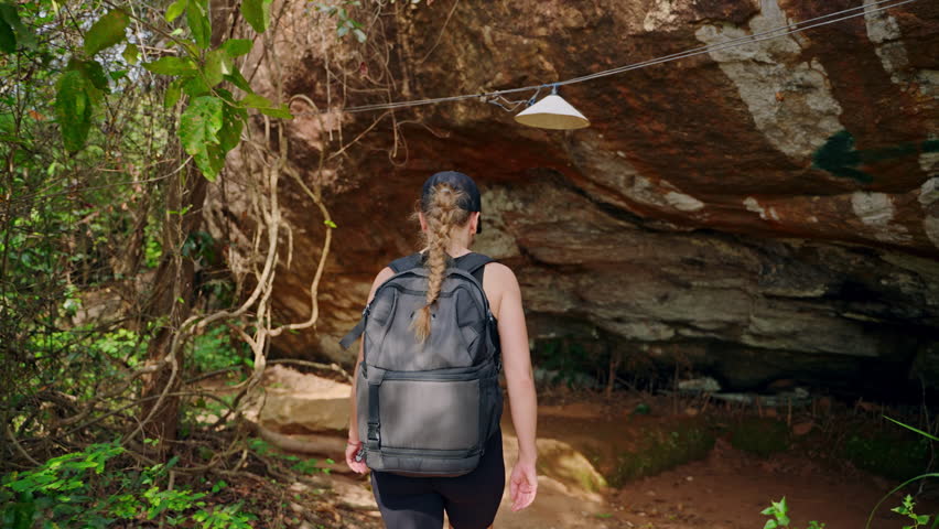 Solo female hiker explores forest grotto on rocky trail. Adventure travel, active lifestyle in nature. Backpacker treks in woods, touches stone, hikes off beaten path near Pidurangala Rock. Slowmo