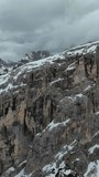 Aerial around view of amazing rocky mountains in snow under moody gray clouds, Dolomites, Italy. Vertical video