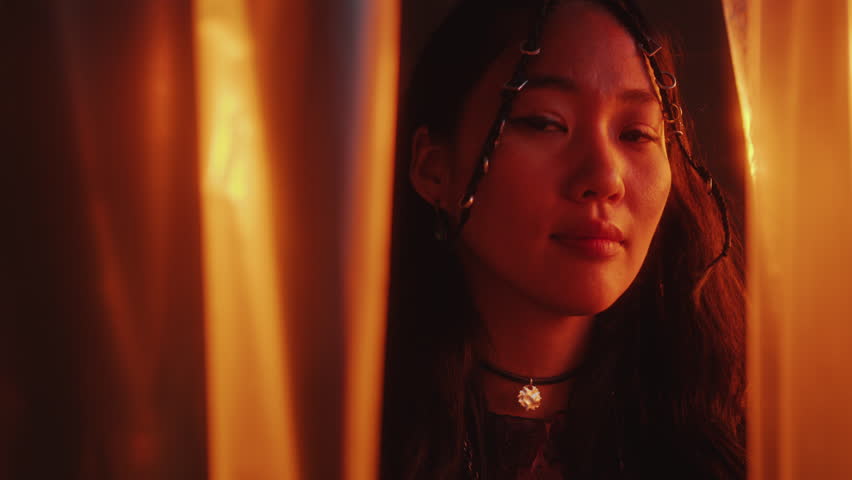 Young beautiful Asian woman with stylish hair braids standing among plastic wrap curtains and posing on camera in studio with multicolored neon light. Video portrait