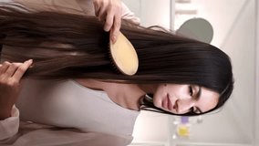 Vertical video. Strands brushing. Feminine routine. Beautiful woman in elegant blouse combing gorgeous healthy smooth long brunette hair in light home interior. - Powered by Shutterstock - Get 15% off with code: PIKWIZARD15