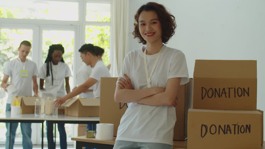 Medium portrait of young female student smiling at camera while volunteering in donation organization with collegemates