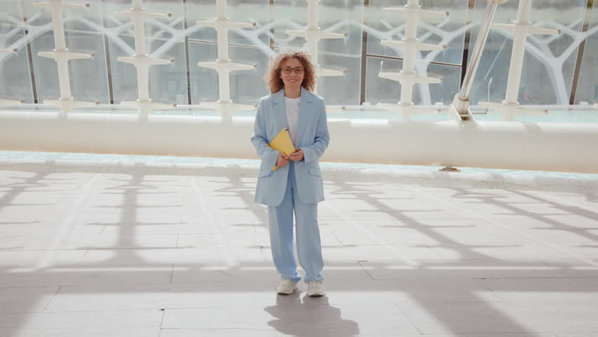 Ebullient young woman with voluminous curly hair twirls in a blue suit, holding a yellow book, with a white geometrically-designed structure as backdrop. Slow motion. 