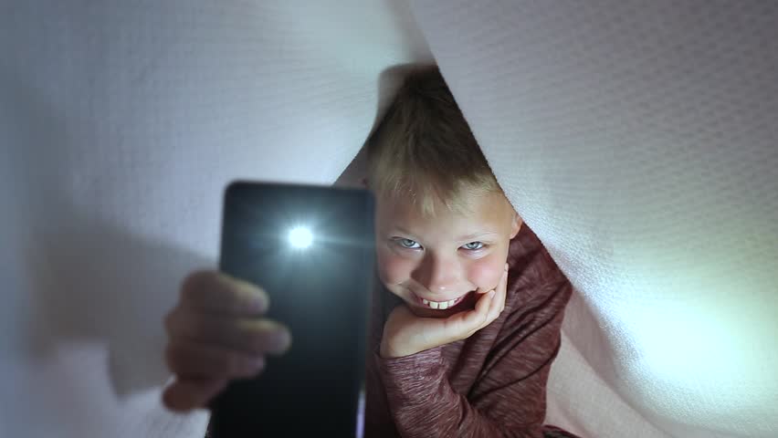 Funny child using flashlight of his smartphone while looking at camera. Boy hiding under white blanket. 