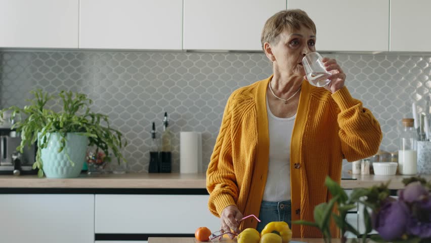 A senior Caucasian woman drinking water standing in the bright kitchen