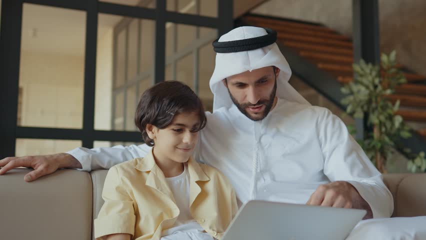 Father and son studying together on the couch. Man from emirati wearing kandura traditional dress teaching to his son on the laptop.