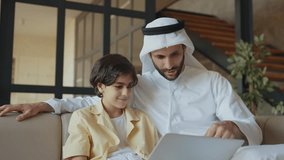 Father and son studying together on the couch. Man from emirati wearing kandura traditional dress teaching to his son on the laptop. - Powered by Shutterstock - Get 15% off with code: PIKWIZARD15