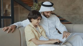 Father and son studying together on the couch. Man from emirati wearing kandura traditional dress teaching to his son on the laptop. - Powered by Shutterstock - Get 15% off with code: PIKWIZARD15