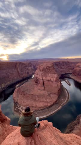 A young adult man open his arms in sign of freedom while sitting in front of Horseshoe Bend while rest and enjoying the landscape view at top of Grand Canyon National Park in Arizona, USA