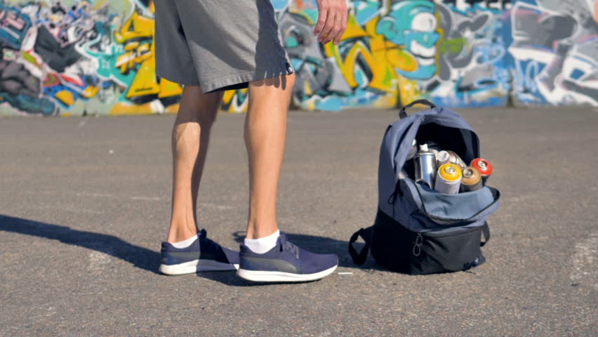A graffitist takes a backpack with paints in one hand.  