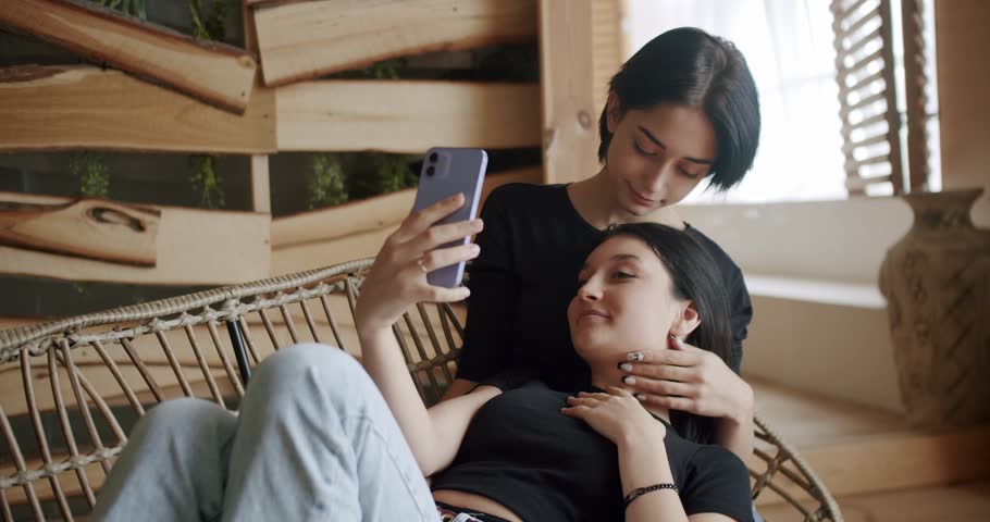 Low angle handheld shot of young lesbian couple smiling and touching hands gently while sitting on sofa against window in light living room at home. Two women sharing love and support holding hands.