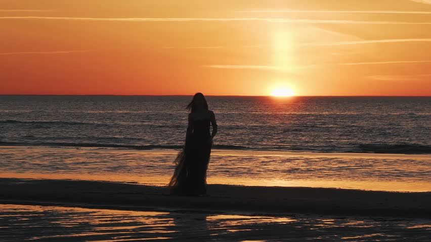 Beautiful lady in a long cocktail dress enjoying magical sunset on the Beach in California. Sunset by the ocean. Romantic concept.