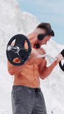 Shirtless man in headphones trains barbell for biceps. Outdoor sports. Vertical video.