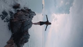 Vertical video woman standing on coastal boulder near ocean, arms open to sea wind. Powerful wind from ocean ruffles hair and dress flutters. Woman enjoys pristine and powerful energy ocean elements.