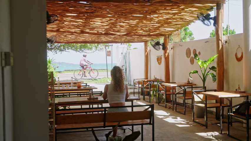 Woman relaxes in tropical cafe, gaze ocean view. Bicyclist passes by idyllic beachside. Casual dining looks serene seascape, offers escape to laid-back lifestyle, summer vibes felt in airy open space.