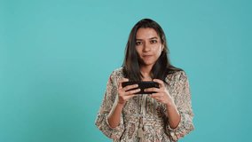 Gamer with disappointed look on face after receiving game over screen, holding smartphone. Indian woman upset after losing videogame, playing with phone, isolated over studio background