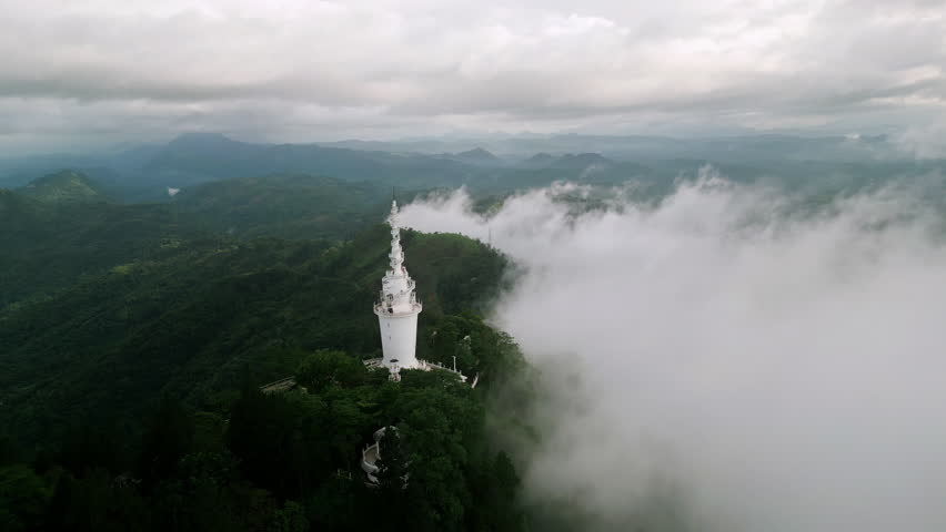 Aerial of drone flies over Ambuluwawa Tower encircled by mist in Sri Lankan mountains. Green plants below, enshrouded monument. Spectacular view for unique travel, natural beauty.