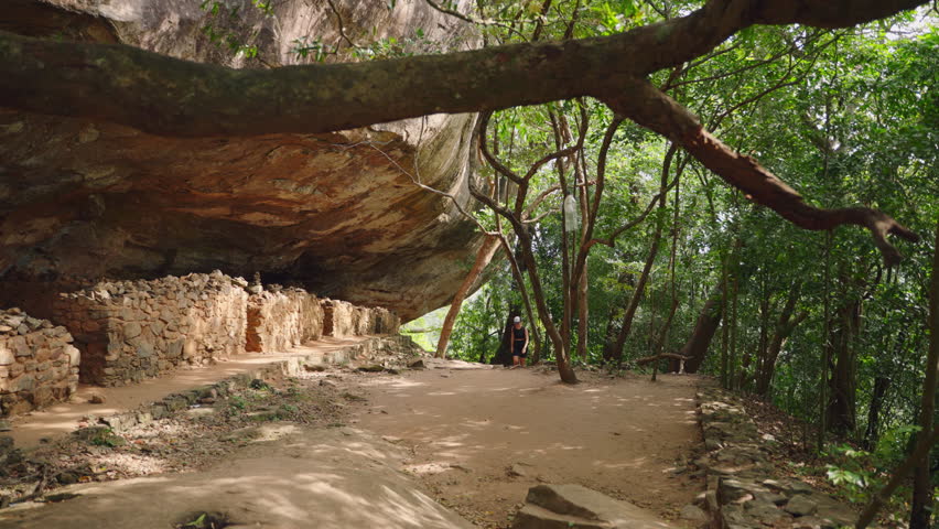 Active woman hikes in mountains, explores ancient grotto trail. Rests, drinks water, enjoys nature. Solo traveler adventure, rocky path, green plants. Healthy lifestyle, outdoor exploration on hike.