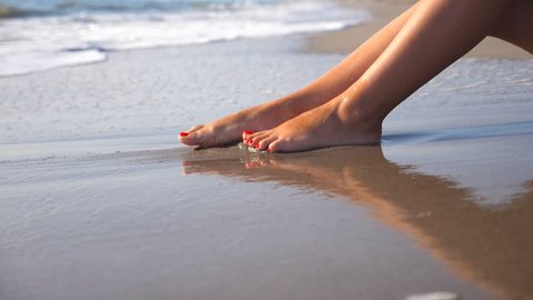 Sea waves washing over tanned female feet. Beautiful young woman relaxing on coast during summer vacation. Legs of girl lying on the beach on a sunny day. Close up Slow motion