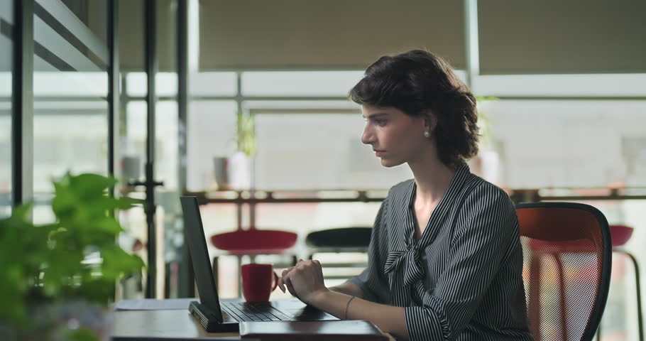 European Caucasian young adult busy woman sitting on chair use laptop indoor office place. Gen z female worker do online project work typing check email hold mobile answering phone call centre job