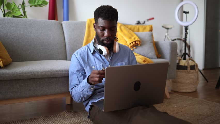 African American professional focuses on an online transaction, using a credit card and laptop from his comfortable home office. Man Processing Online Payment While Working Remotely