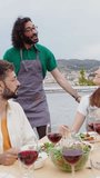 Vertical HD video of young group of diverse people toasting red wine at summer party celebration. Multiracial friends sitting around dinner table while enjoying bbq dinner together at terrace.