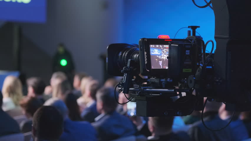 A detailed view of a professional video camera capturing a live event with a crowd of attendees in a conference hall. Focus on technology and event documentation.