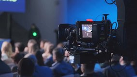 A detailed view of a professional video camera capturing a live event with a crowd of attendees in a conference hall. Focus on technology and event documentation.