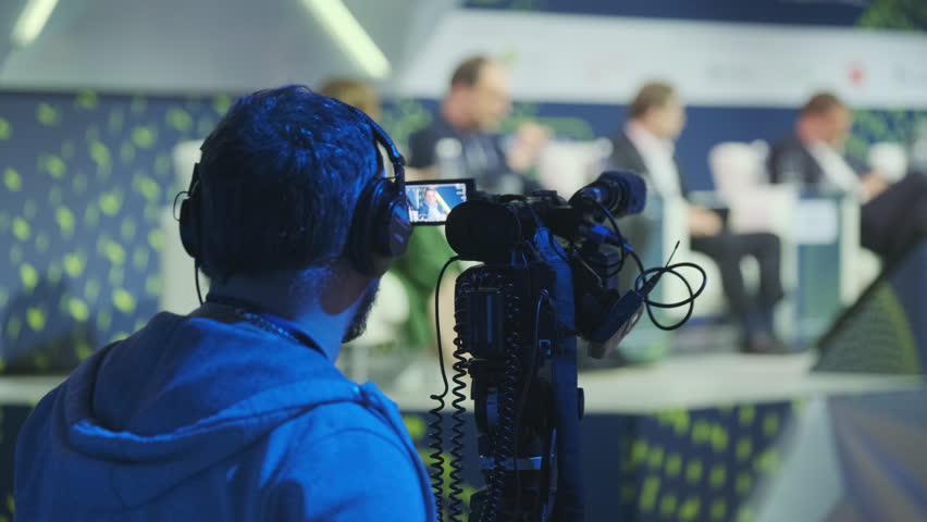A professional videographer captures a business conference on camera, focusing on speakers discussing from the stage, highlighting the importance of media coverage in corporate events.
