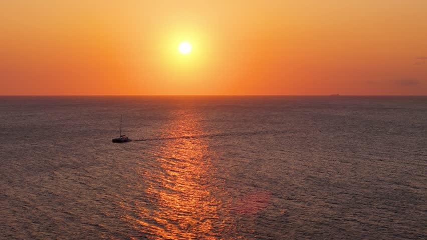 Panoramic aerial tracking follows catamaran silhouette from side as sun glows orange sky
