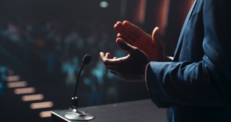 Close Up of an Anonymous Conference Speaker Applauding on Stage Behind a Podium Stand. Person Showing Approval and Praising the Event by Clapping. Audience is Cheering in the Background