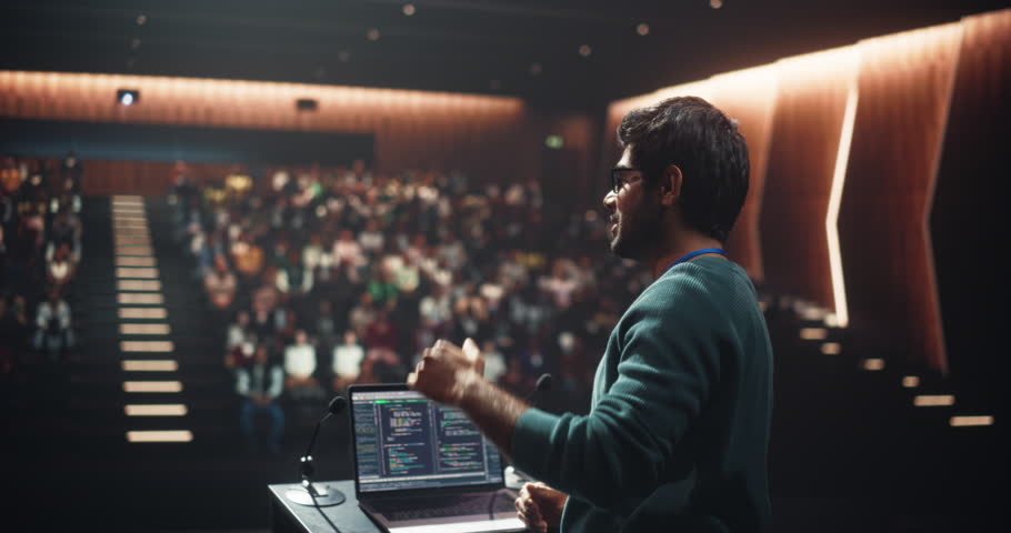 Software Company Lead Developer Making an Inspiring Presentation About Innovation at Technology Conference. Young Indian Man Using Laptop Computer and Talking to an Audience in a Dark Hall