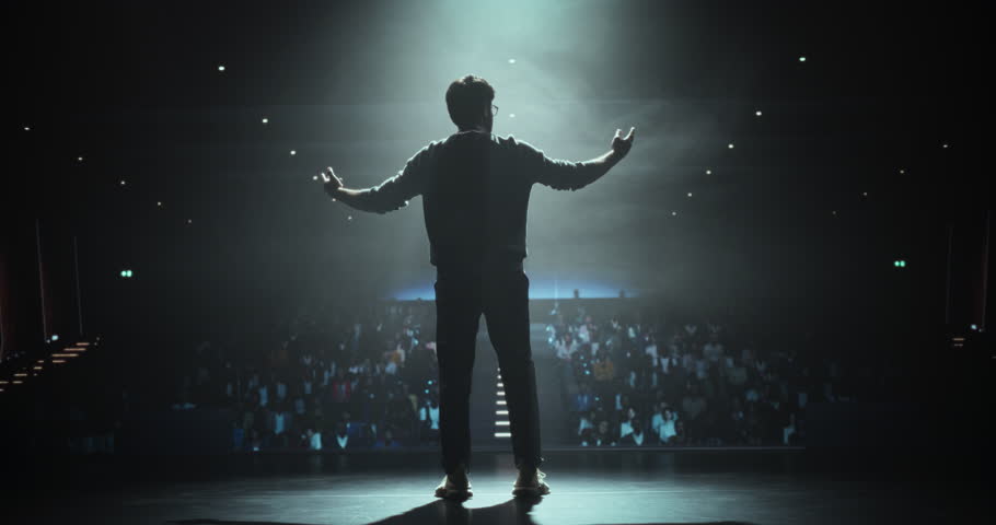 Cinematic Footage of an Anonymous Man Standing Under a Spotlight on Stage in Front of a Sold-Out Audience. Motivational Speaker or a Stand Up Comedian Making an Unforgettable Performance