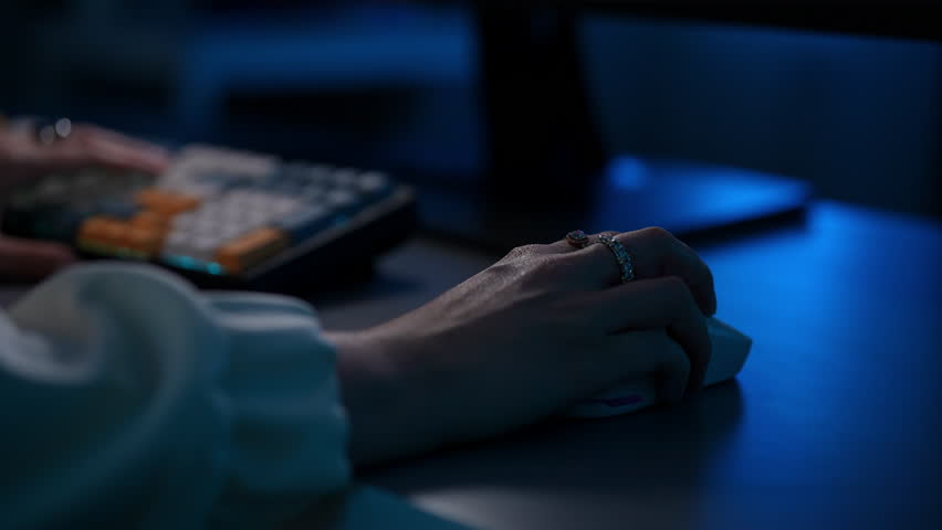 Closeup hands of female gamer during video game on pc desktop on dark futuristic workspace. Close-up side view of freelancer programmer typing on wireless mechanical keyboard and using mouse.