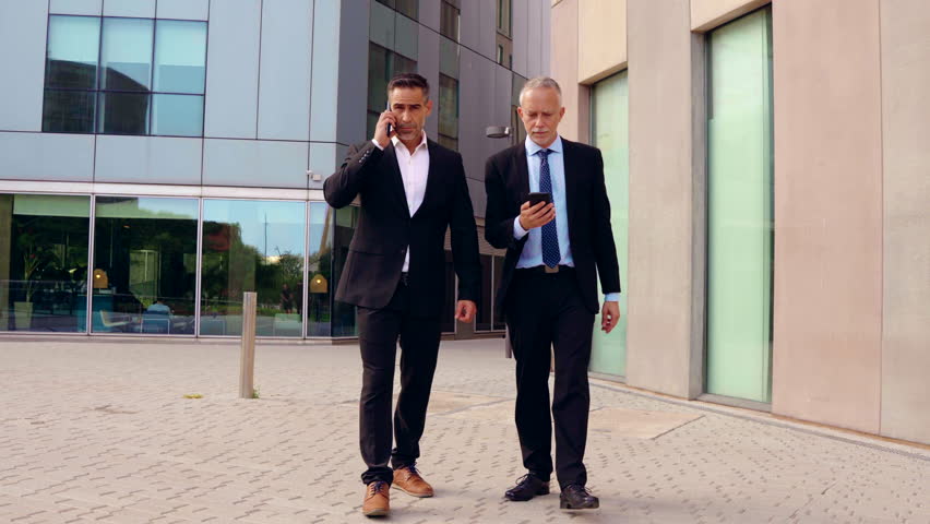 Two attractive and self-confident businessmen walking with smartphones outdoors.