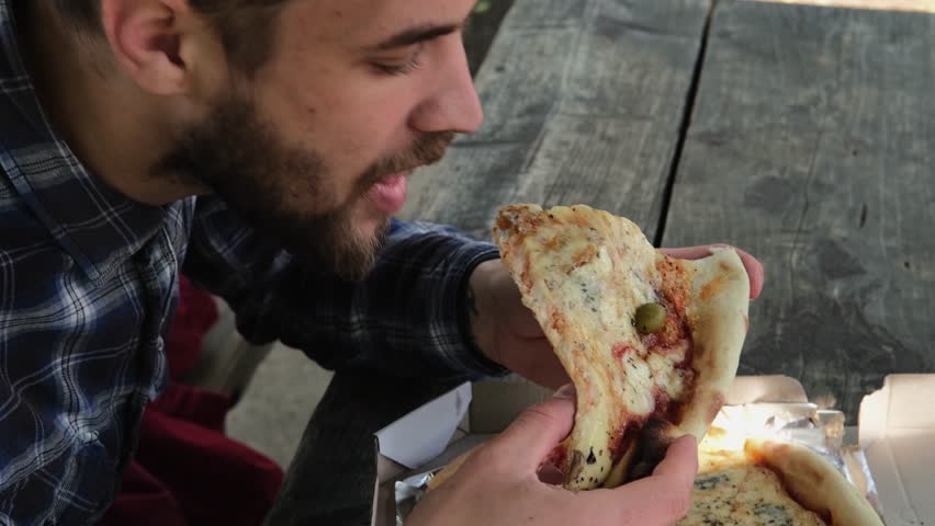 Gen z, food and hunger of young person enjoying carbs pizza slice meal with satisfied face. A young Caucasian man is sitting in a park and eating cheese pizza, enjoying the taste of food alone.