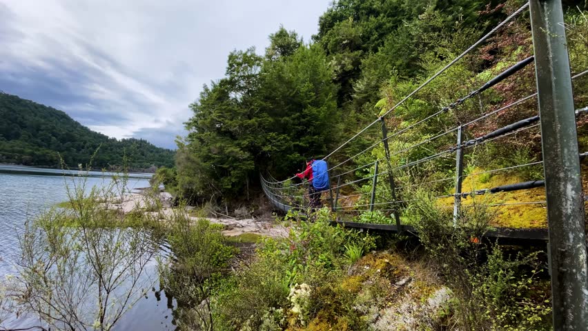 A hiker with a backpack walks across a suspension bridge in the wilderness next to a lake