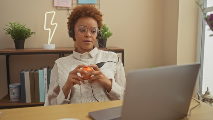 African american woman working from home with laptop, headphones, and stress ball in a cozy room setting. Royalty-Free Stock Footage #3502145339