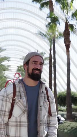 Vertical. Handsome Caucasian man strolling smiling in Valencia city science park. Cheerful young tourist walking lively on sunny winter day. Male looking around happy. Cultural tourism millennial