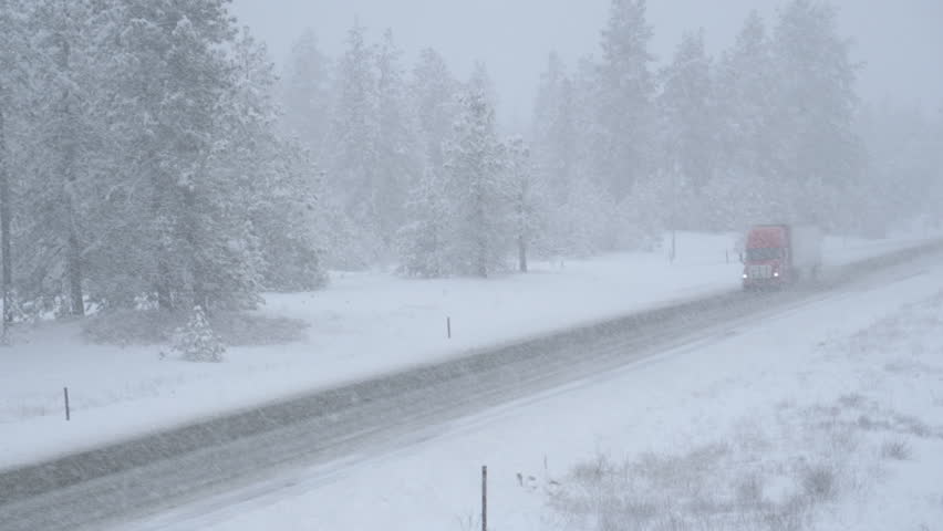Red semi truck hauls a heavy cargo container across the state of Washington and through a snowstorm. Freight lorry navigates the slippery country road in the low visibility during an intense blizzard.