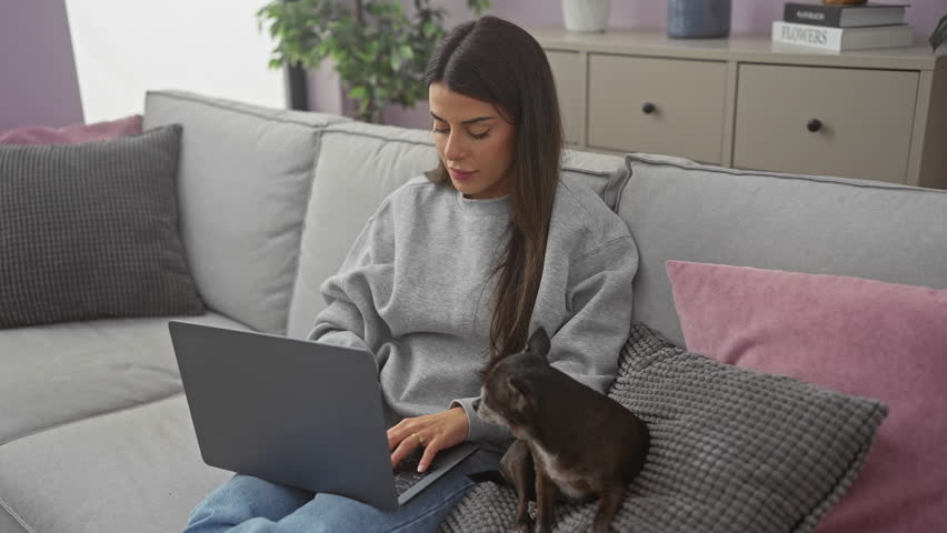 Young woman smiles at camera with arms crossed, sitting with chihuahua on sofa in a cozy apartment room, laptop closed beside her.