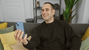 Smiling hispanic man using smartphone for video call in a cozy living room decorated with plants.