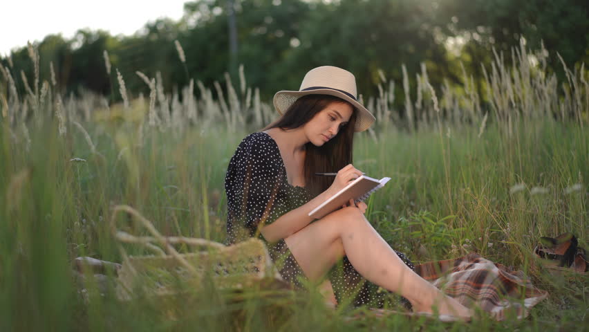 A girl with dark long hair in a brimmed hat writes with a pen in a notebook, sitting on a plaid in tall green grass in a field on a summer sunny day outdoors