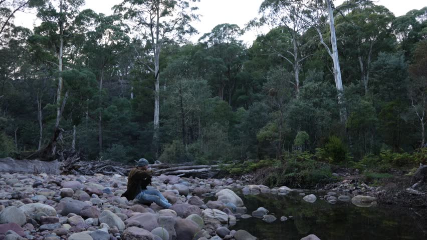 A bushman smoking a pipe by a river in the Australian bush.