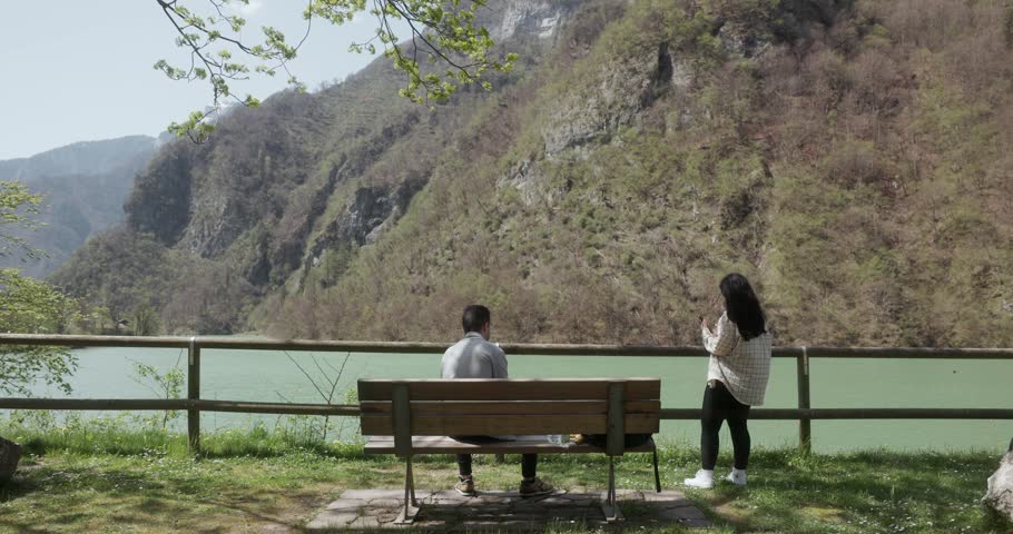 Woman Taking Pictures Of A Man Eating On The Park Bench By The Lake. - wide shot