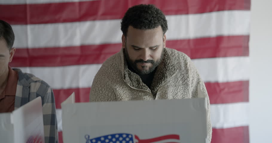 Mixed race man voting at election booth. Camera moves from medium shot to close up and he looks up, into lens. US flag in background. 4K DCI